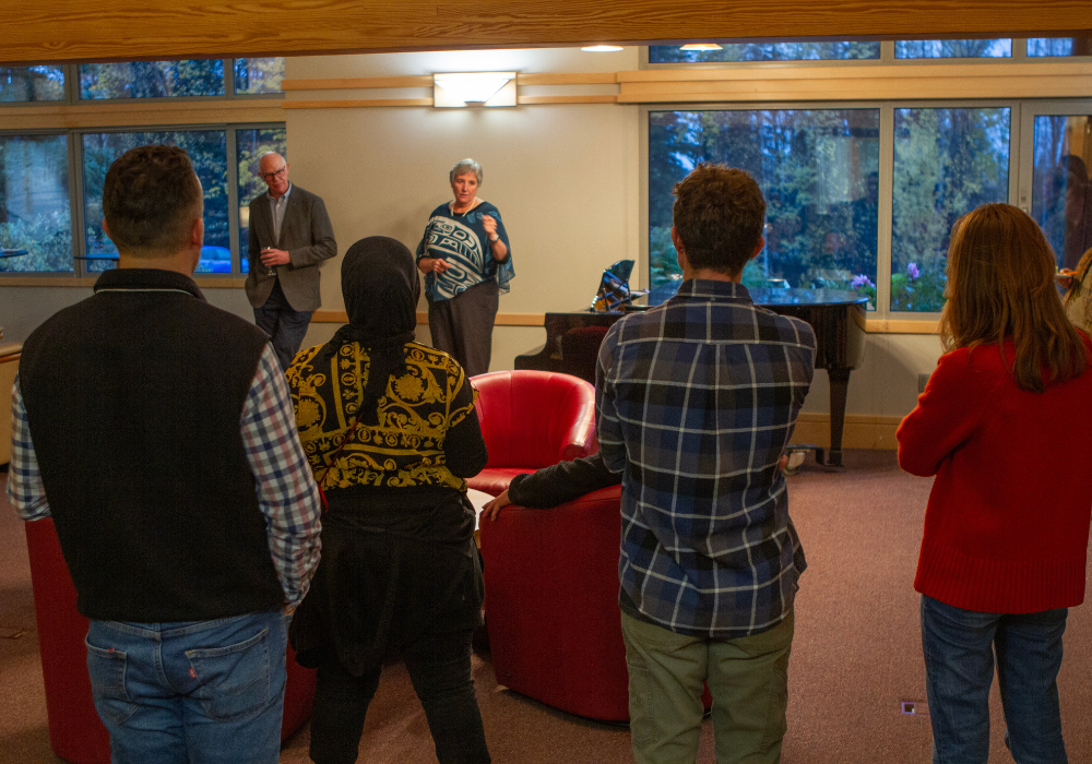 People stand in a warmly lit living room listening to a speaker address the group during a dinner reception at the University of Alaska President’s House.
